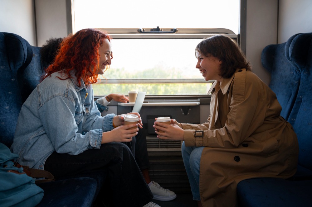 Two women sitting face-to-face in a train compartment, smiling and enjoying hot drinks while engaging in conversation. The scene is bright and relaxed, with natural light coming through the window behind them.